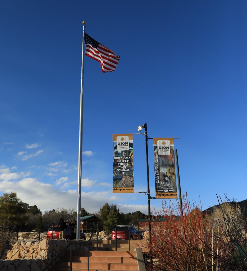 A U.S. flag flies beside informational banners at the entrance of Zion National Park against a blue sky.